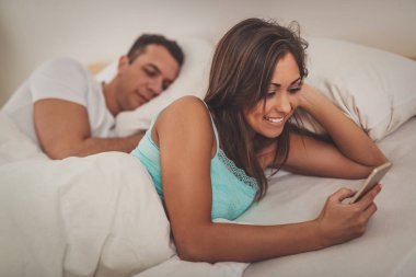 Genç adam gülümseyen kız arkadaşı yatakta akıllı telefon kullanırken uyuyor..