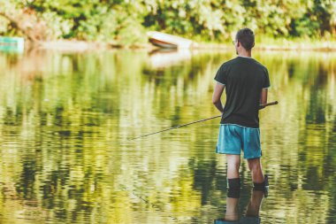 A young man in boots is fishing standing in shallow water freshwater of lake fringed with lush scenery.