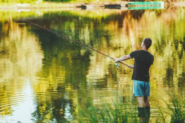A young man in boots is fishing standing in shallow water freshwater of lake fringed with lush scenery 