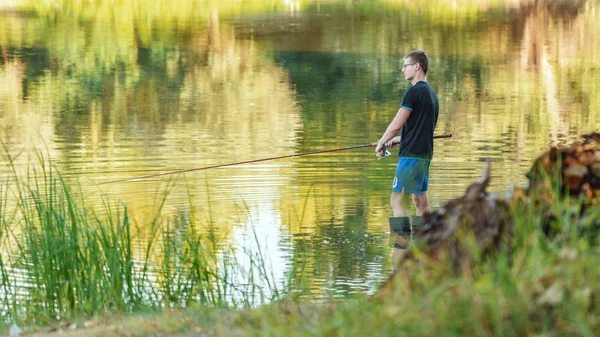 A young man is fishing from a coast of a freshwater lake fringed with lush scenery.