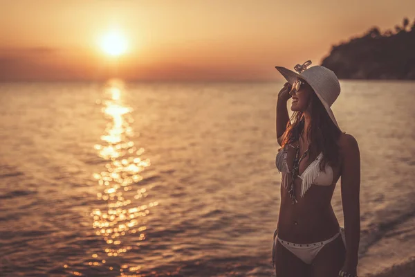 A beautiful woman in white bikini is enjoying in sunset on the beach with  white hat on her head.