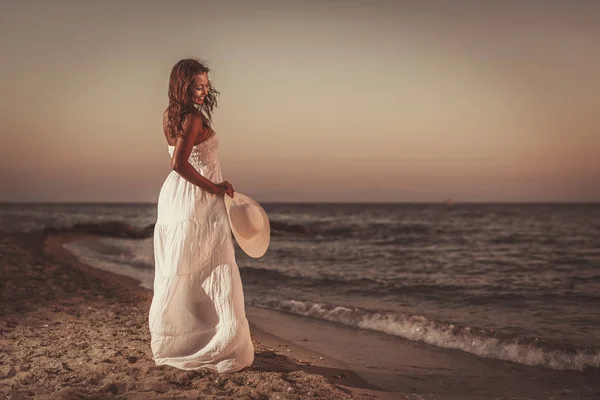 一位身穿白衣的漂亮女人正在海滩上享受日落时分 还戴着白帽 — 图库照片