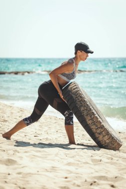 Orta yaşlı, kaslı bir kadın. Plajda antrenman yaparken lastik çeviriyor. 