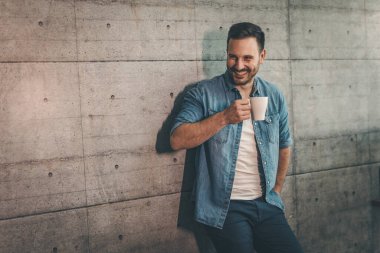 Genç başarılı girişimci bir fincan kahveyle beton duvarın önünde duruyor.