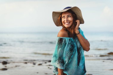 Güneşli bir günde güzel genç bir kadın sahilde dinleniyor. Poz veriyor ve yüzünde tebessümle uzaklara bakıyor.. 