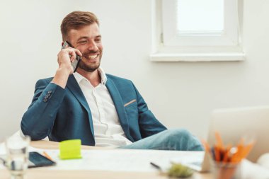 Genç başarılı mimar, ofiste yeni bir projenin planları üzerinde çalışırken akıllı telefondan konuşuyor.. 