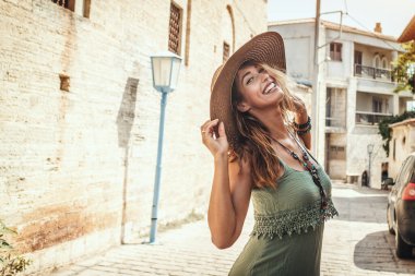 Güzel, gülümseyen genç bir kadın Akdeniz 'de bir kasabanın sokaklarında yürüyor ve güneşli yaz günlerinin tadını çıkarıyor. Poz veriyor ve yüzünde hasır şapka ve gülümsemeyle kameraya bakıyor..