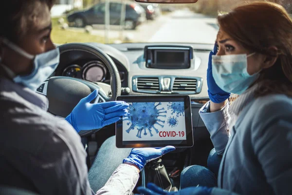 Young business women with protective masks on their faces are sitting in a car and talking about global crisis. They are showing each other an animation of a corona virus on a tablet.