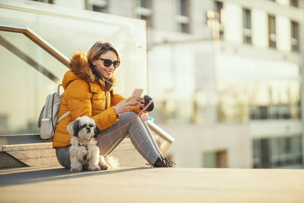 Beautiful young fashion woman is spending time with her cute pet dog playing in the city street looking at the smartphone.