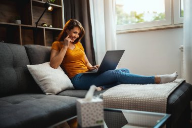 Çekici genç bir kadın kanepede oturuyor ve bilgisayarını evde kullanıyor..
