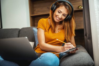 Sevimli genç kız öğrenci kanepede oturuyor, dizüstü bilgisayarını ve kulaklıklarını kullanarak internetten ders izliyor ve evden öğreniyor. Genç bir kadın, öğretmenini takip ederken video çağrısını not alıyor.. 