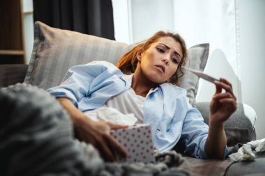 Genç kadın hasta yatarken kanepede termometreye bakıyor..