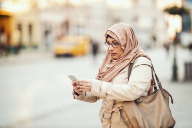 Orta yaşlı, neşeli yüzlü, tesettürlü Müslüman bir kadın şehir ortamında dikiliyor, akıllı telefonundan mesajlar yolluyor..