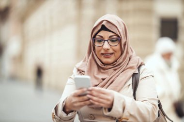 Orta yaşlı, neşeli yüzlü, tesettürlü Müslüman bir kadın şehir ortamında dikiliyor, akıllı telefonundan mesajlar yolluyor..