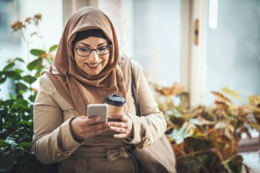 Mutlu yüzlü tesettürlü orta yaşlı Müslüman kadın pencerenin kenarında duruyor ve akıllı telefonuna mesajlar yolluyor..