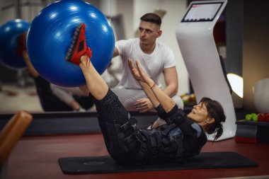 Gülümseyen son sınıf öğrencisi bir kadın spor salonunda kişisel antrenörüyle oturma egzersizleri yapıyor. Siyah bir takım giyip kaslarını canlandırmak için EMS elektronik simülatörü kullanıyor..