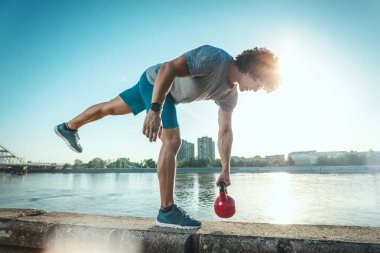 Vücut geliştirici güçlü bir vücut çalışması yapıyor. Duvarda kettlebell var. Nehrin kenarında, mavi gökyüzüne karşı..