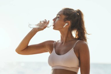 Güzel genç bir kadın yaz güneşli bir günde deniz sahilinde antrenman yaparken dinleniyor ve su içiyor..
