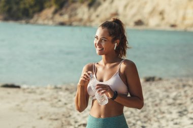 Güzel genç bir kadın yaz güneşli bir günde deniz sahilinde antrenman yaparken dinleniyor ve su içiyor..