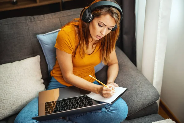 Cute young female student sitting on the sofa, using her laptop and headphones to watching lesson online and learning from home. Young woman taking notes while following teacher doing lesson on video call.