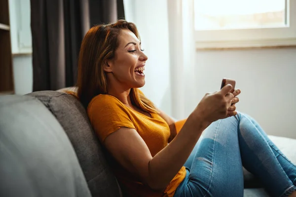 매력적 여성이 소파에 앉아서 스마트폰으로 집에서 비디오 게임을 — 스톡 사진