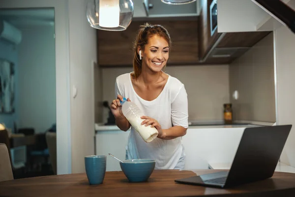 Beautiful young woman is preparing her healthy breakfast in her kitchen and using laptop to make a video chat with someone.