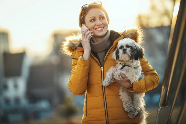 美しい若いファッションの女性は 彼女のかわいいペット犬と一緒に街の通りで時間を過ごしているとスマートフォンで話しています — ストック写真