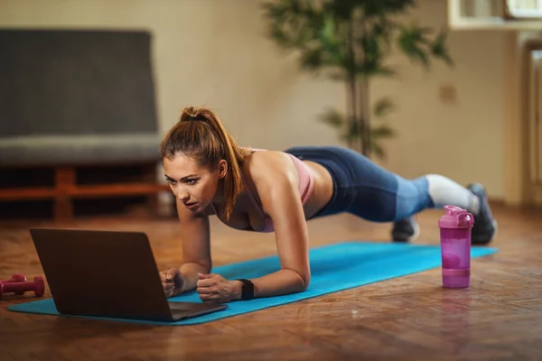 Young smiling woman is doing plank exercises in the living room on floor mat at home, looking at the laptop, in morning sunshine.