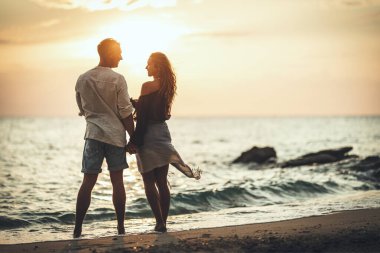 Sevgi dolu bir çift gün batımında kumlu kumsalda eğleniyor ve sarılıyor. Birbirlerine bakıyor ve mutlu bir şekilde gülümsüyorlar..