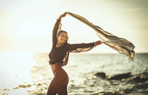 一个年轻貌美的女人在日落时正在海滩上玩得很开心 也很放松 她双手拿着透明的围巾 摆好姿势 望着别处 — 图库照片