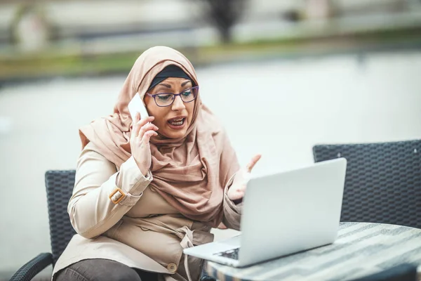 Middle aged worried Muslim woman wearing hijab is sitting on the cafe in urban environment, talking by smartphone and working on laptop.