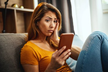 Endişeli genç bir kadın kanepede oturuyor ve evde sosyal medyada sörf yapmak için akıllı telefonu kullanıyor..