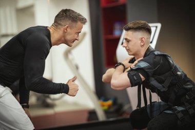 Genç bir adam spor salonunda kişisel antrenörüyle çömelme egzersizleri yapıyor. Siyah bir takım giyip kaslarını canlandırmak için EMS elektronik simülatörü kullanıyor..