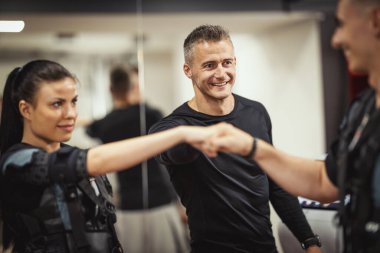 Siyah takım elbiseli, elektromanyetik simülatörlü güzel bir çift spor salonunda spor yaptıktan sonra antrenörlerini kutluyorlar..