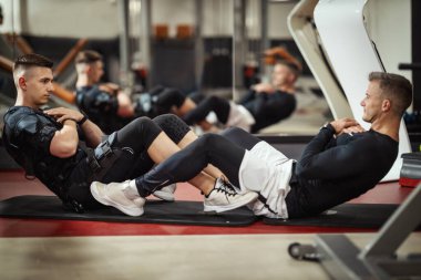 Genç bir adam spor salonunda kişisel antrenörüyle oturma egzersizleri yapıyor. Siyah bir takım giyip kaslarını canlandırmak için EMS elektronik simülatörü kullanıyor..