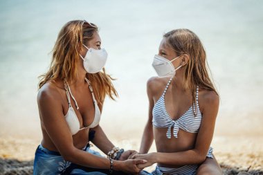Korumalı N95 maskeli, gülümseyen bir kız kardeşin Corona Salgınında sahilde vakit geçirmesi. Doğada kendilerini izole ediyorlar..