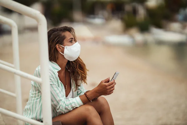 スマートフォンを使って保護マスクをした魅力的な幸せな若い女性のショットとCovid 19中にビーチで休暇を楽しむ — ストック写真