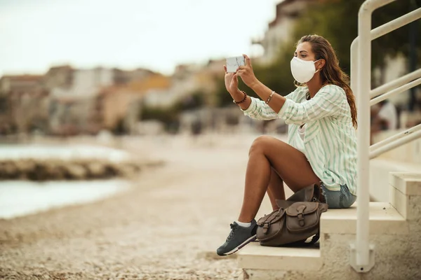 魅力的な幸せな若い女性のショット保護N95マスクを着用し Covid 19中にビーチで休暇を楽しんでいる間に彼女のスマートフォンで写真を撮る — ストック写真