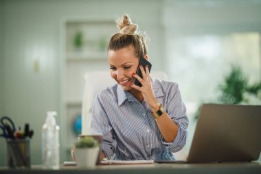Çekici genç bir kadının COVID-19 salgını sırasında bilgisayarında çalışırken akıllı telefon kullanması..