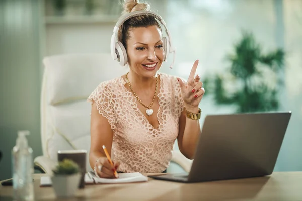 Shot of a attractive young businesswoman sitting alone in her home office with headphones and working on laptop during COVID-19 pandemic.