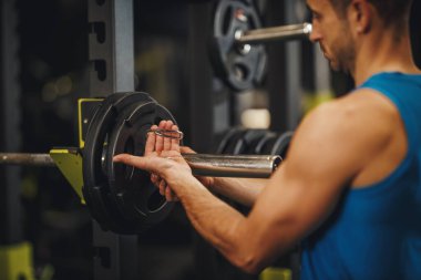 Spor giyim bölümünde spor salonunda çalışan kaslı bir adamın fotoğrafı. Bir sonraki egzersiz serisine başlamadan önce halterlere daha fazla ağırlık koyuyor..