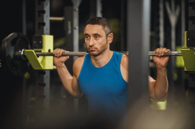 Spor giyimli kaslı bir adamı spor salonunda çalışırken çekmişler. Halterle çömelme egzersizleri yapıyor..