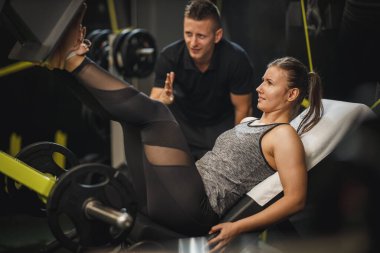 Spor giyimli kaslı genç bir kadını spor salonunda kişisel antrenörle çalışırken çekmişler. Bacak pres makinesinde bacakları için egzersiz yapıyor..