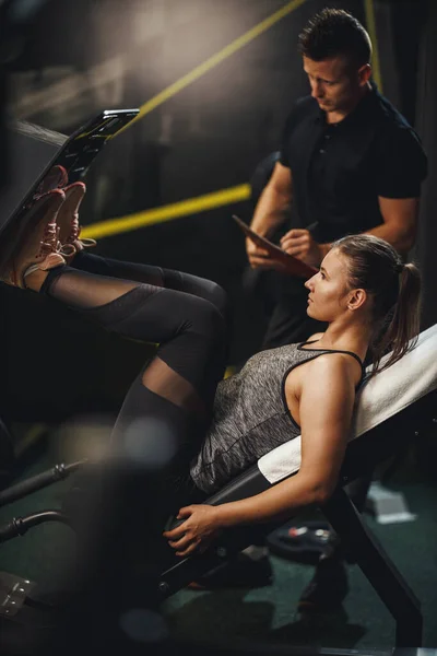 在体育馆和私人教练一起锻炼时被一名身穿运动服的肌肉发达的年轻女子开枪击中 她正在用腿压机为腿做运动 — 图库照片