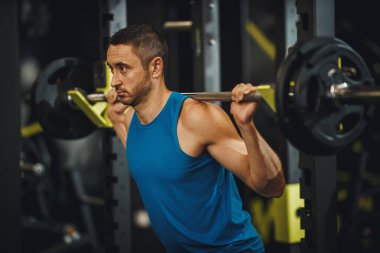 Spor giyimli kaslı bir adamı spor salonunda çalışırken çekmişler. Halterle çömelme egzersizleri yapıyor..