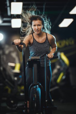 Genç, kaslı bir kadın spor salonunda bisiklet egzersizi yapıyor..