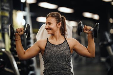 Spor giyimli, kaslı genç bir kadının spor salonundaki sıkı antrenmanında vurulması. O, omuz kaslarını ağır ağırlıkla şişiriyor..