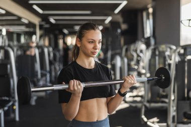 Teen girl doing biceps with barbell in modern gym.