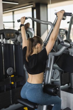 Back view of gym girl in blue legging and black top doing back exercise on machine in gym. 
