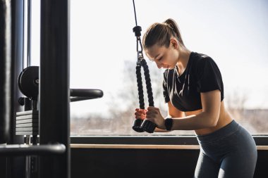 Focused young woman doing triceps on cable machine in modern gym. 